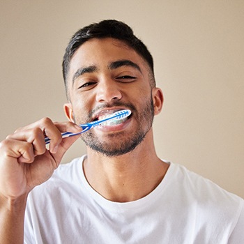 Man in white t-shirt brushing teeth with white and blue toothbrush with tan background